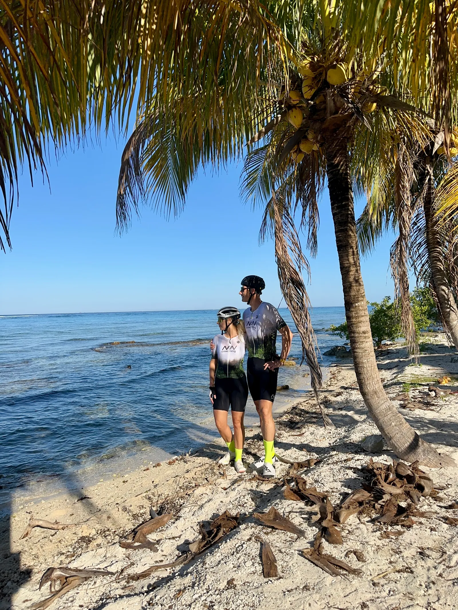 Two cyclists standing beneath palms on a rocky Cozumel shoreline